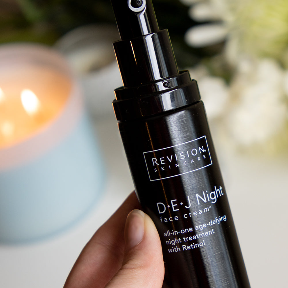 Hand holding a Revision Skincare D.E.J Night face cream bottle with a blurred candle in the background.