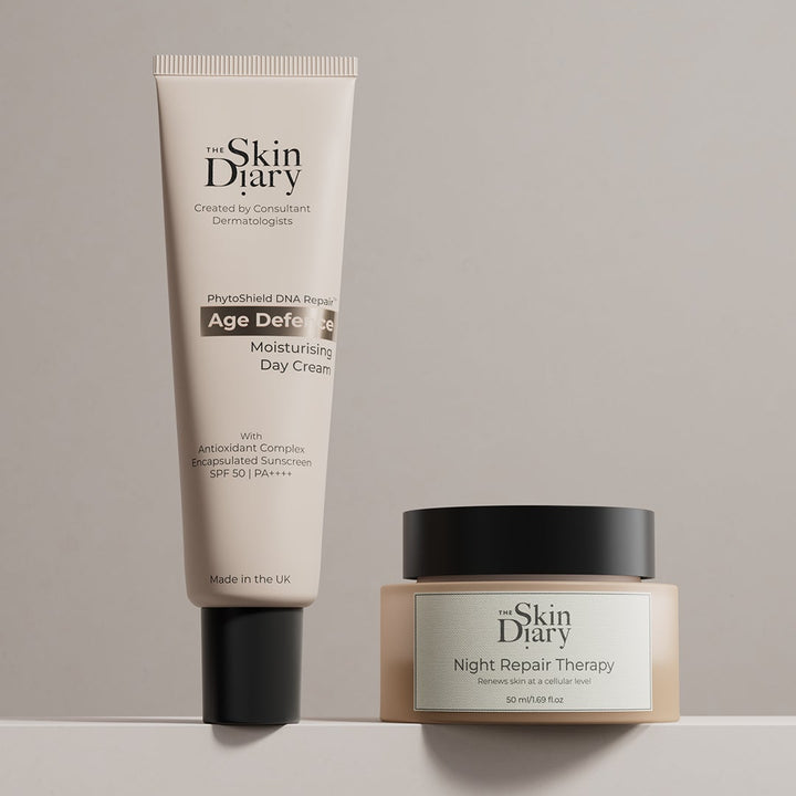 Two skincare products from The Skin Diary, 'Age Defying Day Cream' in a beige squeeze tube and 'Night Repair Therapy' in a beige jar with a black lid, displayed on a light gray surface against a neutral gray background.