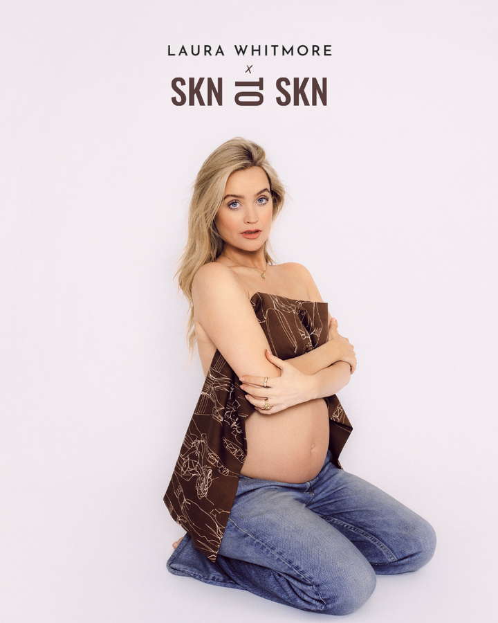 Pregnant woman with blonde hair wearing blue jeans holding a brown patterned cloth over her bare torso under Skn to Skn Laura Whitmore branding